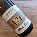 A close-up of a wine bottle labeled 'Riesling Cuvée Albert 2020' by Albert Mann, resting on a wooden surface. The label features a cream background with a central abstract painting in yellow, black, and gray, and the brand name 'Albert Mann' in black script.