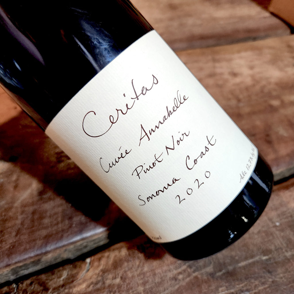 A close-up of a dark wine bottle lying on a rustic wooden surface, showing a cream-colored label with handwritten-style text that reads 'Ceritas Cuvée Annebelle Pinot Noir Sonoma Coast 2020'.