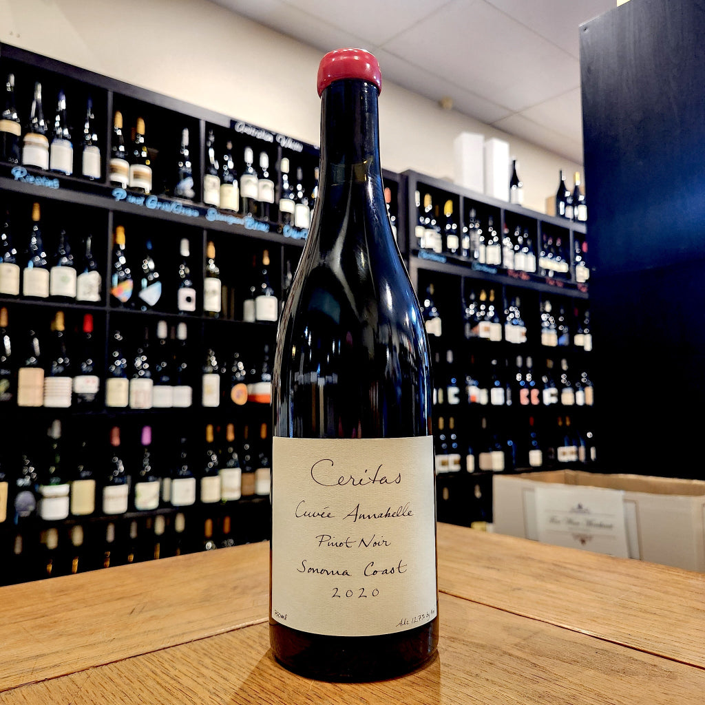 A bottle of 2020 Ceritas Cuvee Annabelle Pinot Noir from the Sonoma Coast standing on a wooden counter in front of a wine cellar shelf filled with bottles.