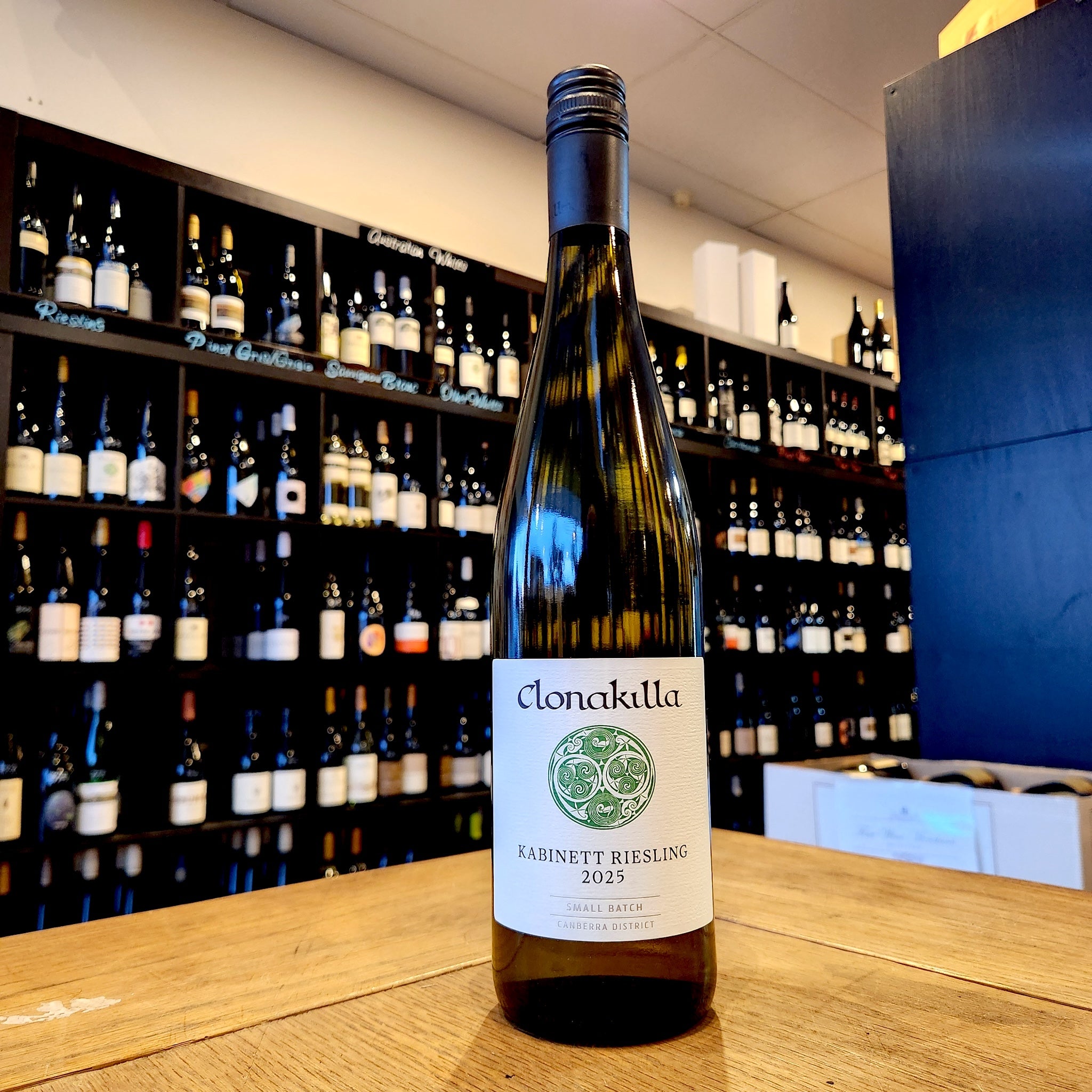 A bottle of Clonakilla Kabinett Riesling 2025 white wine stands on a wooden table in front of a wine cellar shelf filled with bottles.