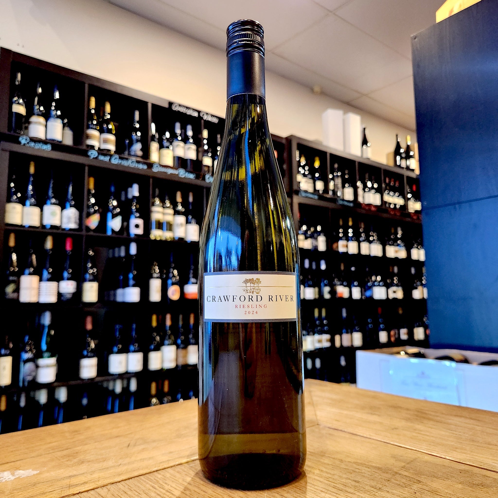 A bottle of Crawford River Riesling 2024 white wine standing on a wooden counter in front of a wine store shelf filled with bottles.
