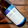 A bottle of Grosset Polish Hill Riesling 2023, angled diagonally on a rustic wooden surface. The label features a white background with blue and gold accents.
