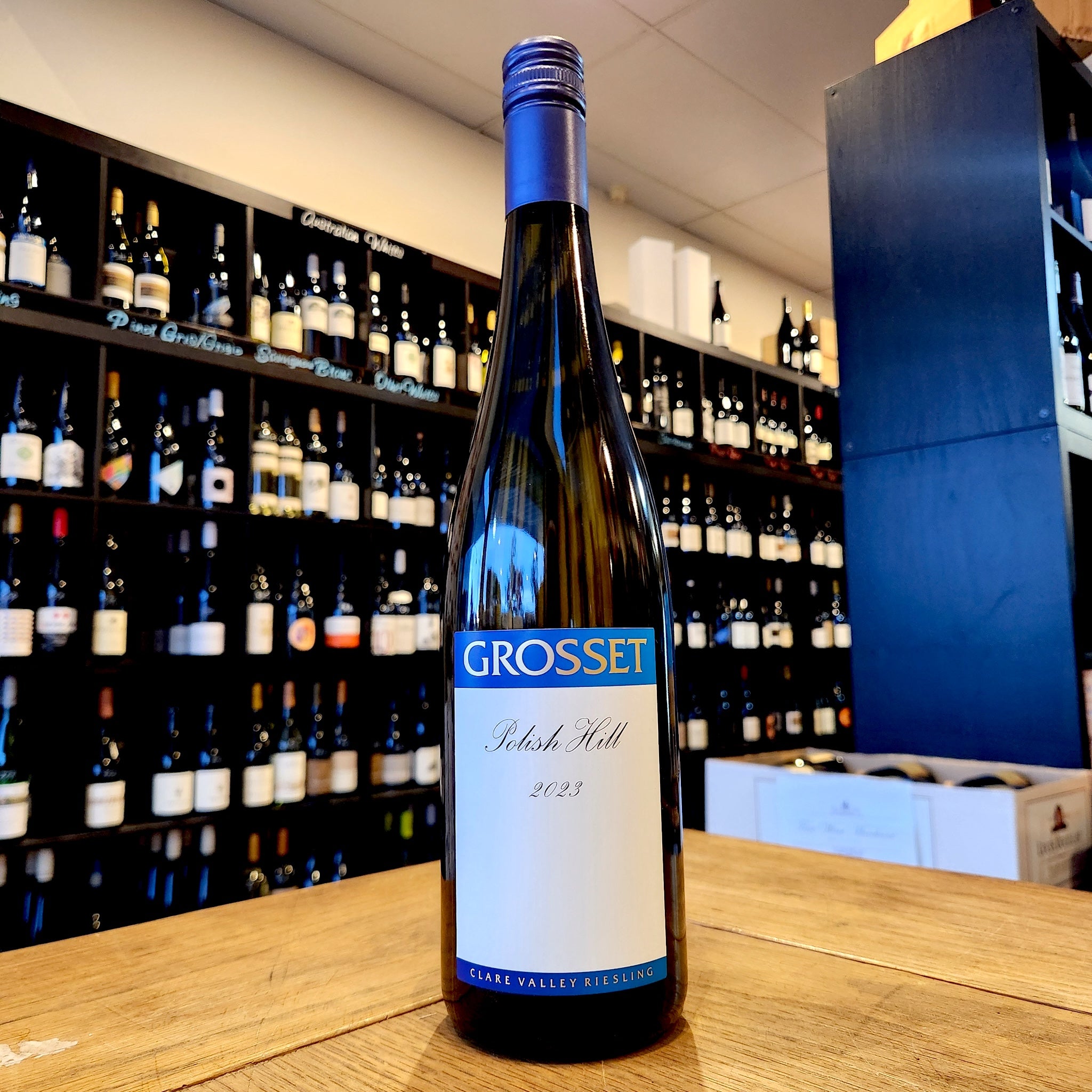 A bottle of Grosset Polish Hill Riesling 2023 white wine stands on a wooden table in front of a wine cellar shelf filled with bottles.