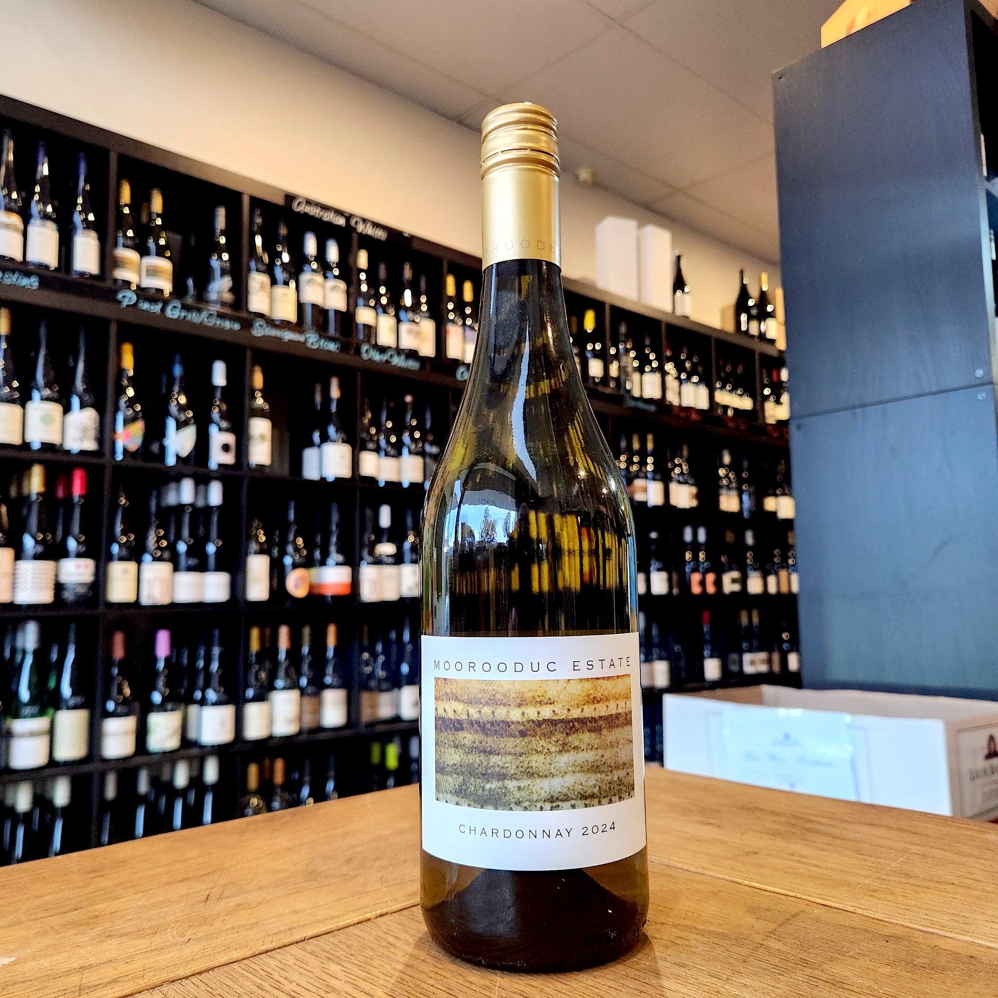 A bottle of Moorooduc Estate Chardonnay 2024 standing on a wooden counter in a wine shop, with shelves of wine bottles blurred in the background.