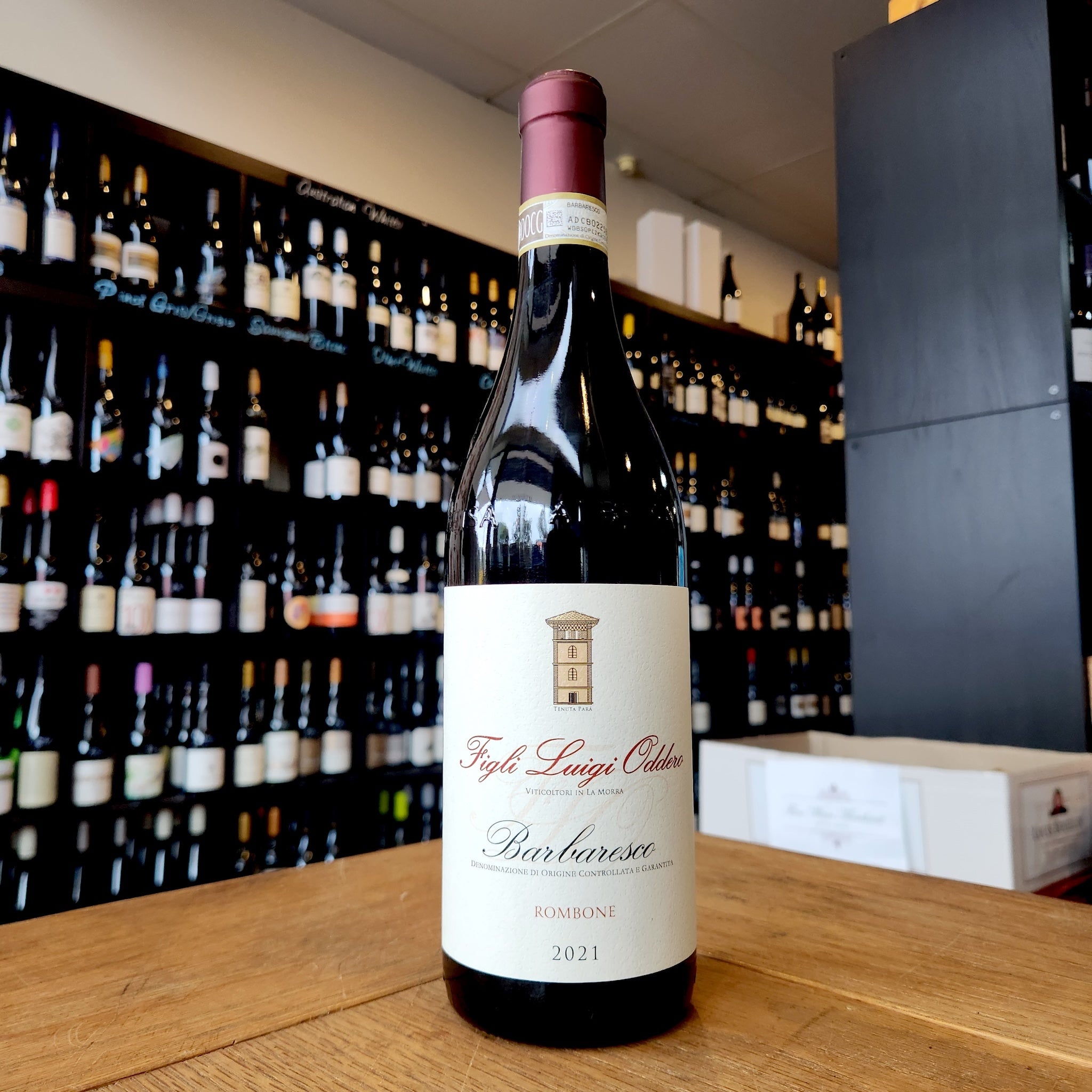 A bottle of 2021 Figli Luigi Oddero 'Rombone' Barbaresco sits on a wooden table in a wine shop with blurred bottles on a shelf in the background.