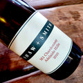 A close-up of a dark glass wine bottle lying on a rustic wooden surface, showing a white label with gold text that reads Shaw and Smith, M3 Chardonnay, Adelaide Hills, 2025.