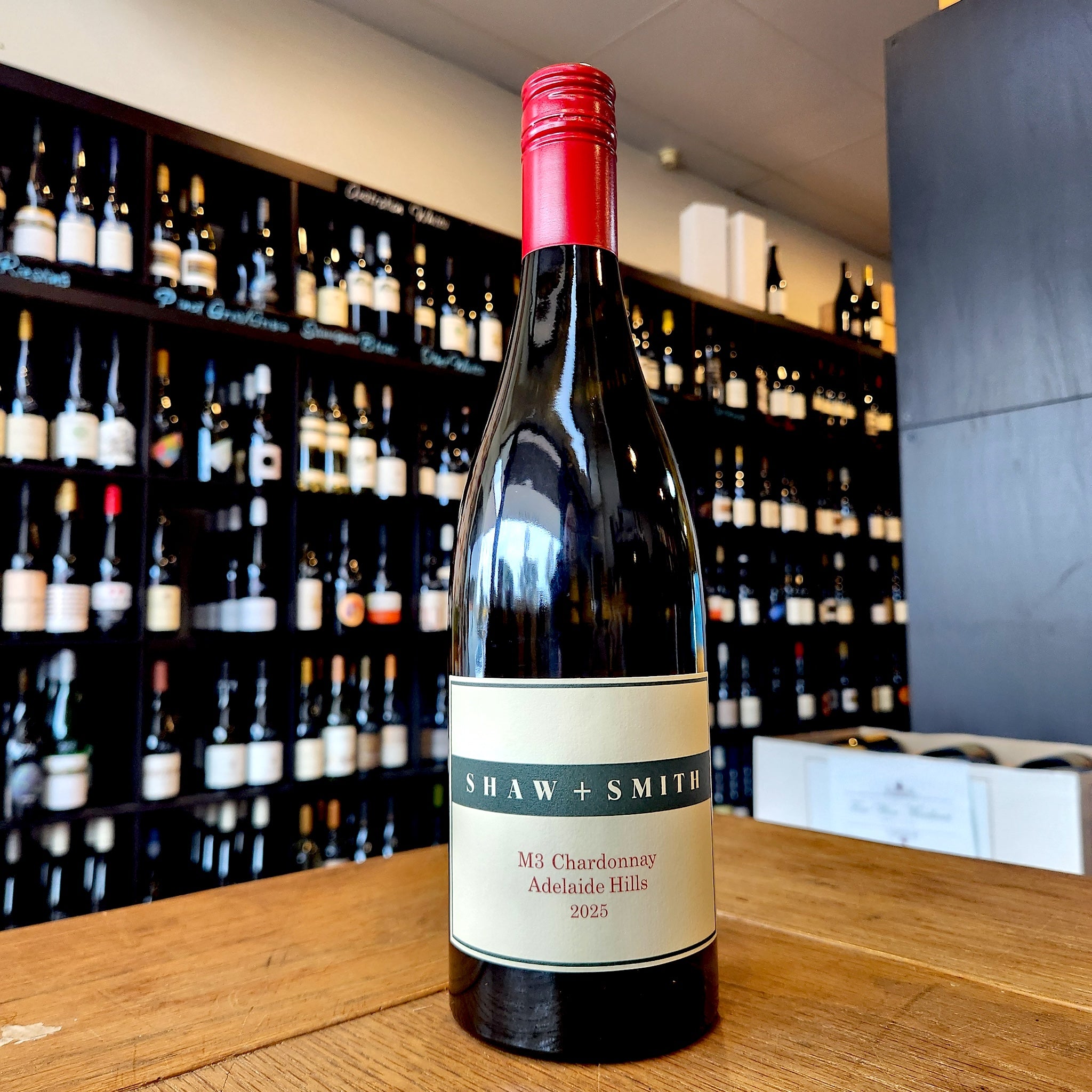 A bottle of Shaw + Smith M3 Chardonnay Adelaide Hills 2025 standing on a wooden table in front of a wine cellar shelf filled with bottles.