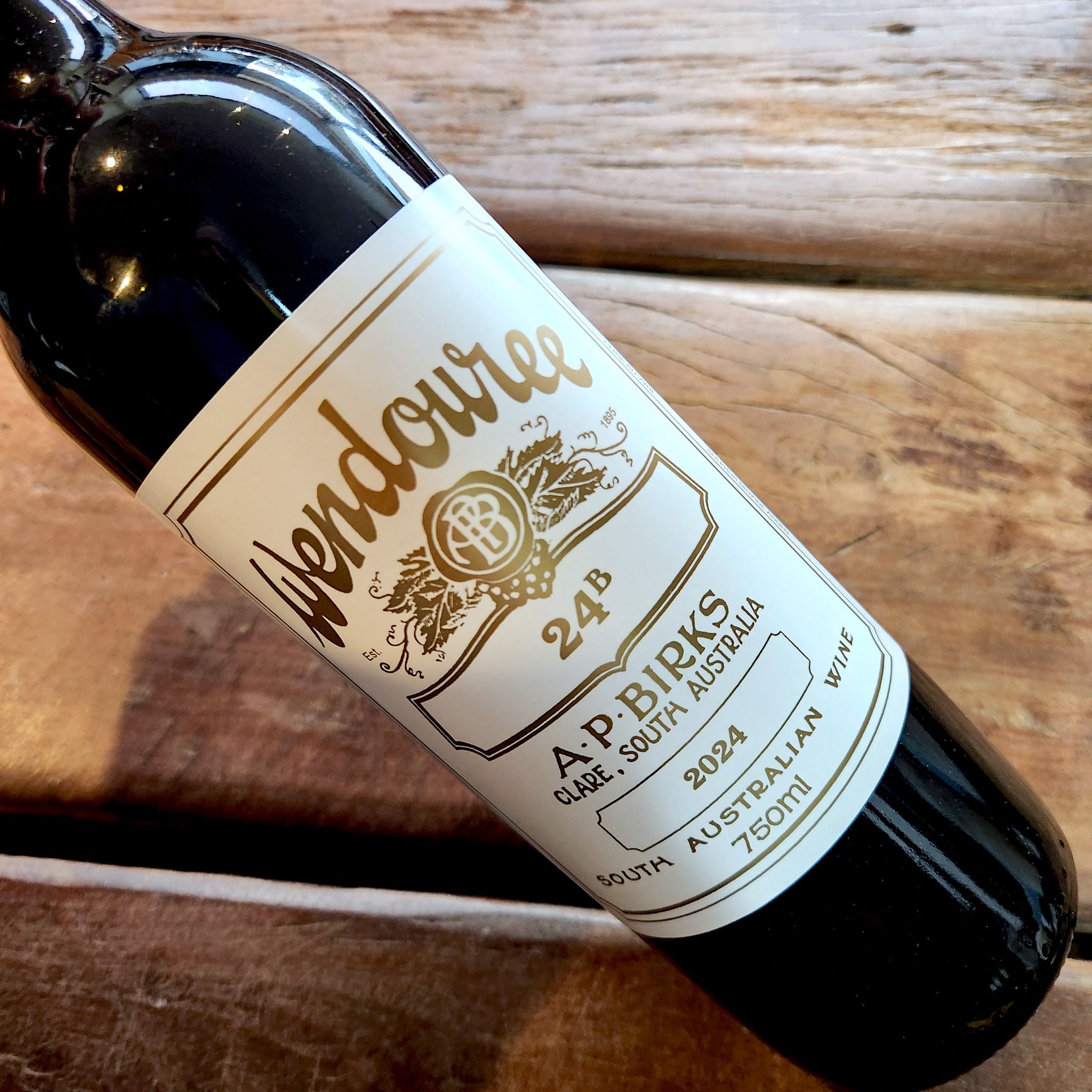 A close-up of a dark glass wine bottle lying on a rustic wooden surface, showing a white label with gold text that reads Wendouree 24B, 2024.