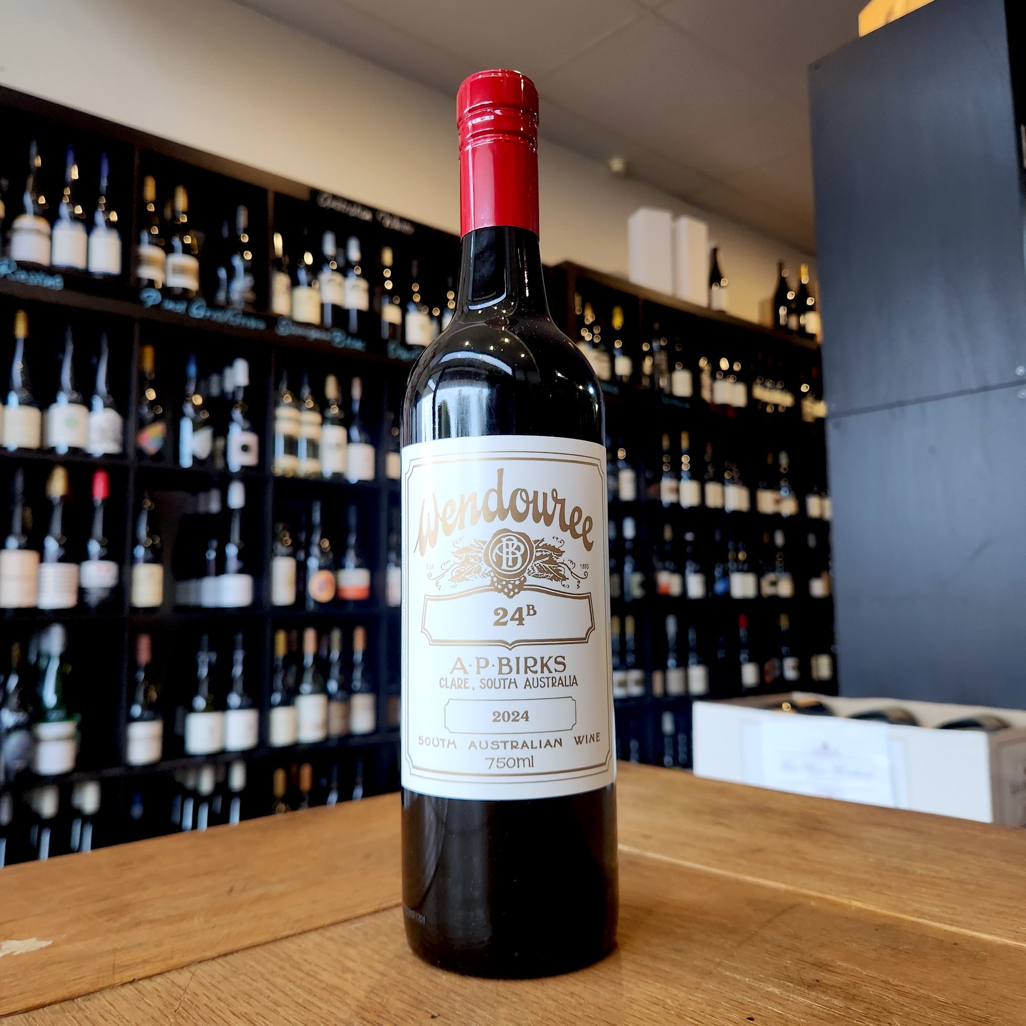 A bottle of Wendouree 24B standing on a wooden table in front of a wine shop wall filled with blurred bottles.
