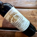 A close-up of a dark glass wine bottle lying on a rustic wooden surface, showing a white label with gold text that reads Wendouree Cabernet Malbec, 2024.