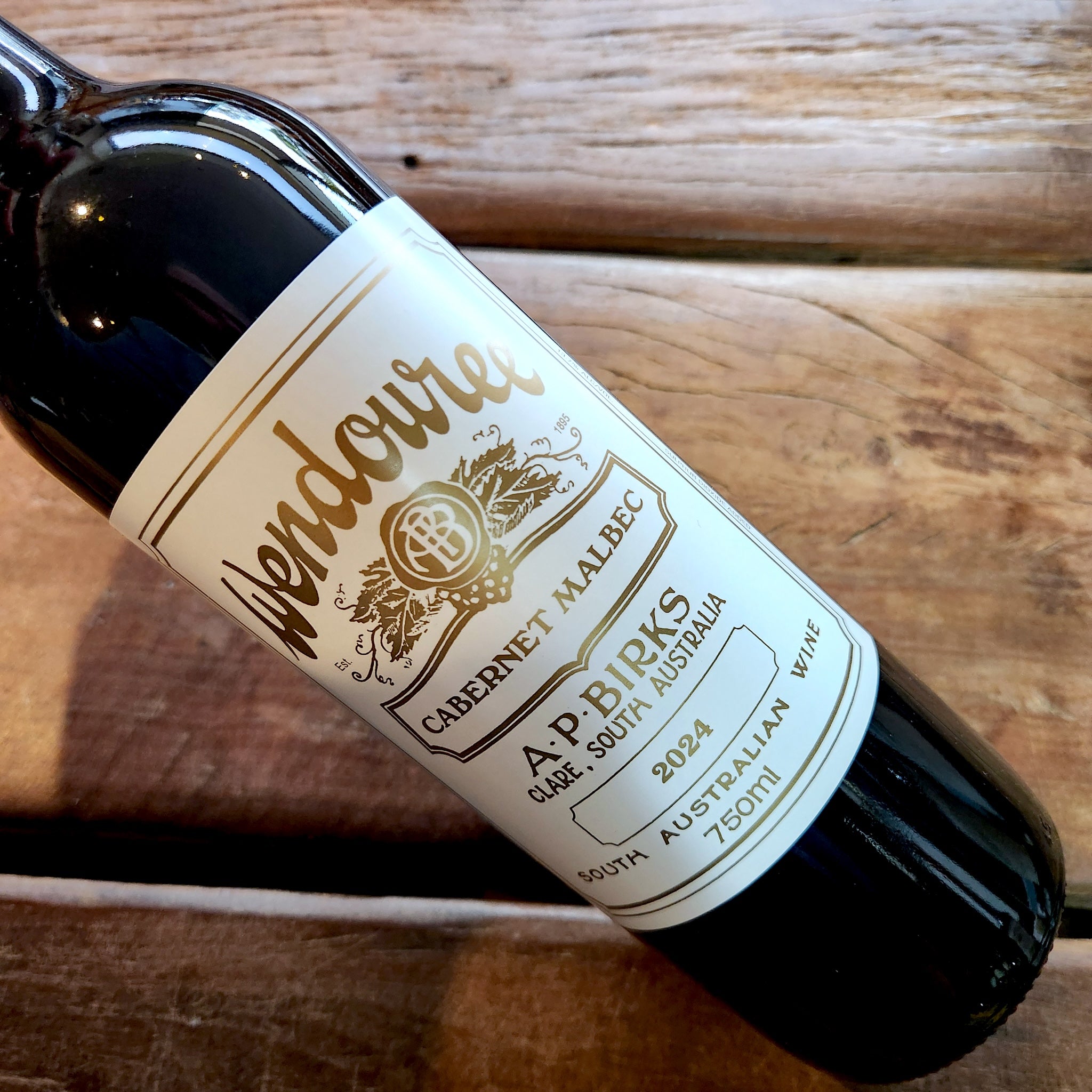 A close-up of a dark glass wine bottle lying on a rustic wooden surface, showing a white label with gold text that reads Wendouree Cabernet Malbec, 2024.