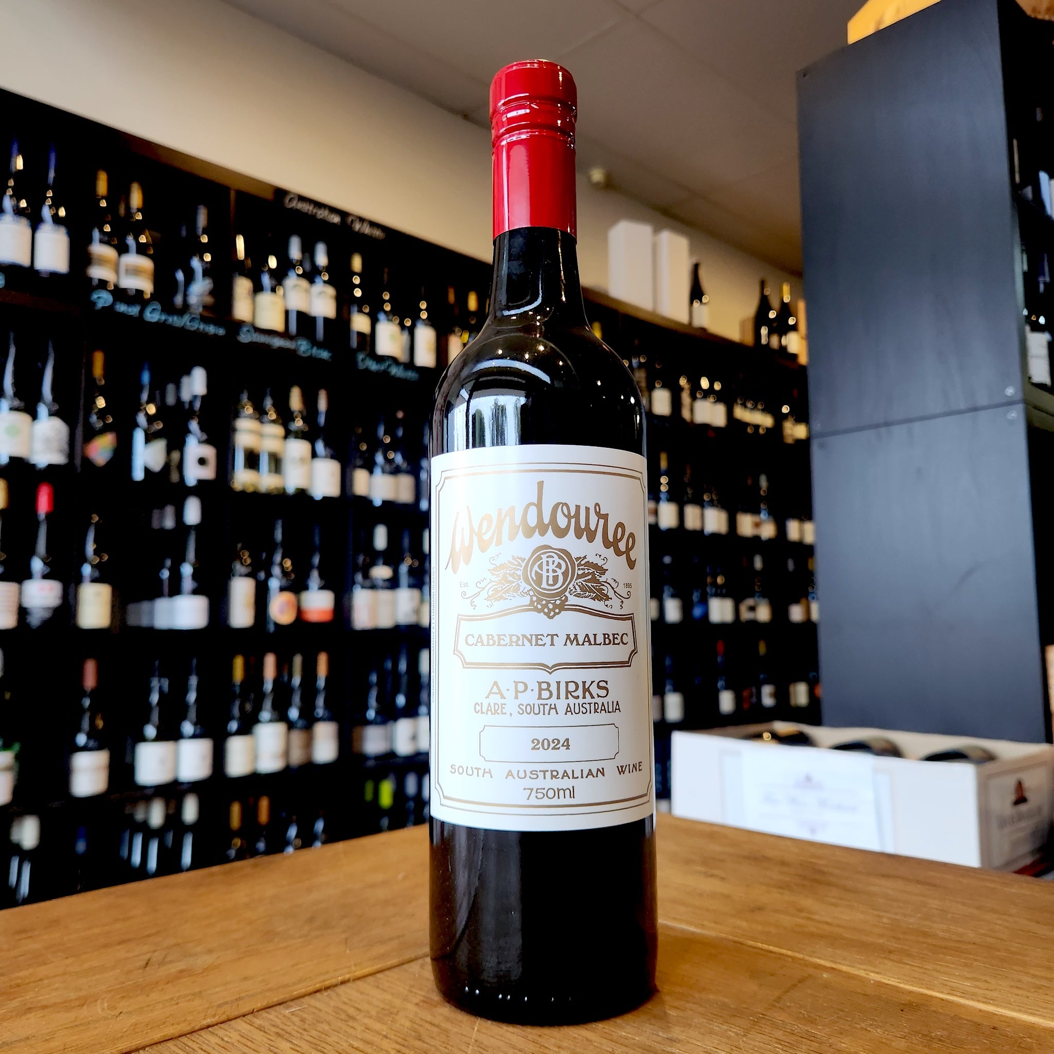 A bottle of Wendouree Cabernet Malbec standing on a wooden table in front of a wine shop wall filled with blurred bottles.