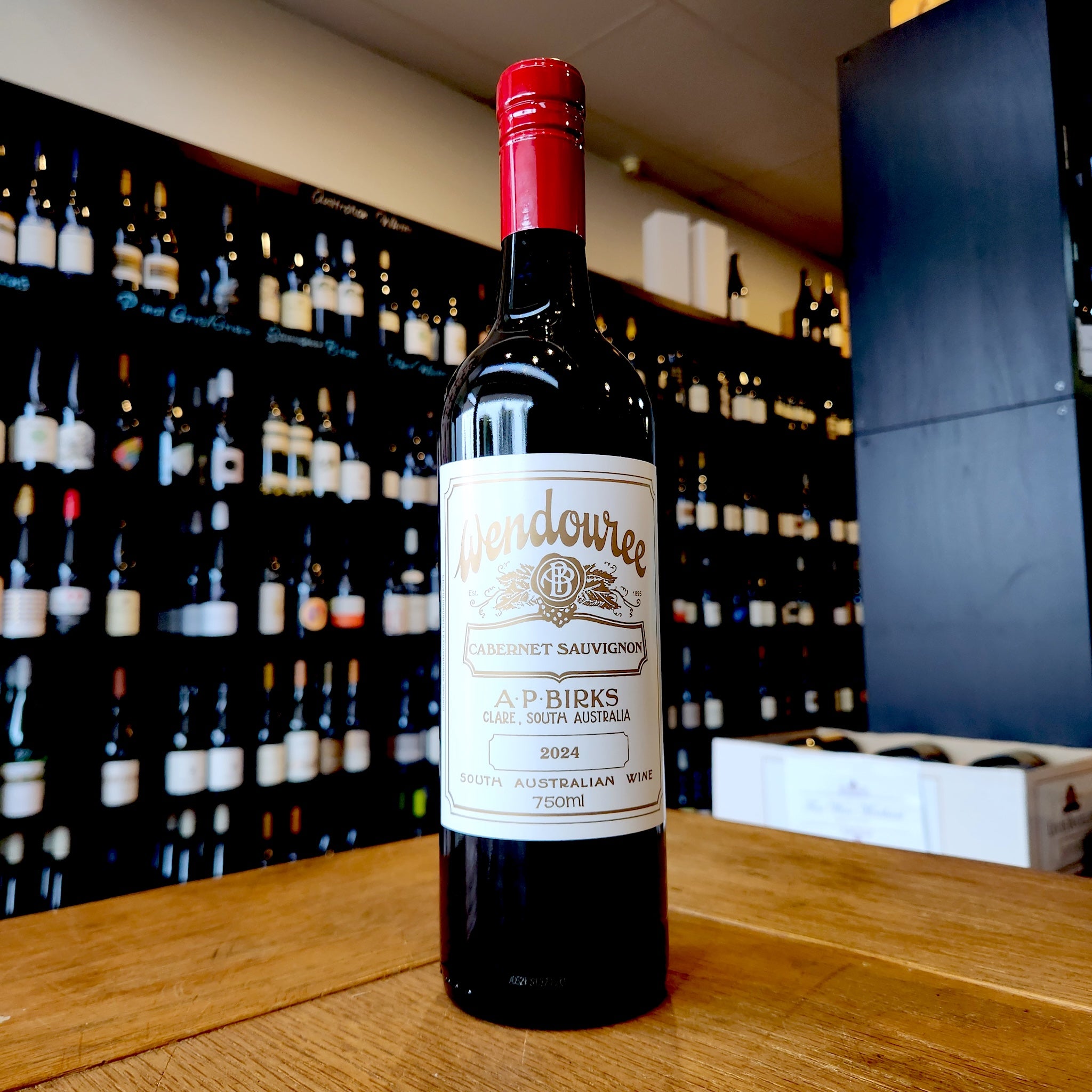 A bottle of Wendouree Cabernet Sauvignon standing on a wooden table in front of a wine shop wall filled with blurred bottles.