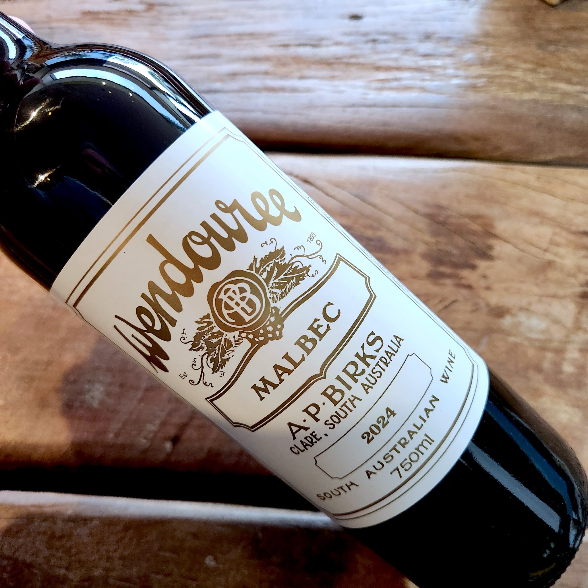 A close-up of a dark glass wine bottle labeled Wendouree Malbec 2024, placed diagonally on a rustic wooden surface.