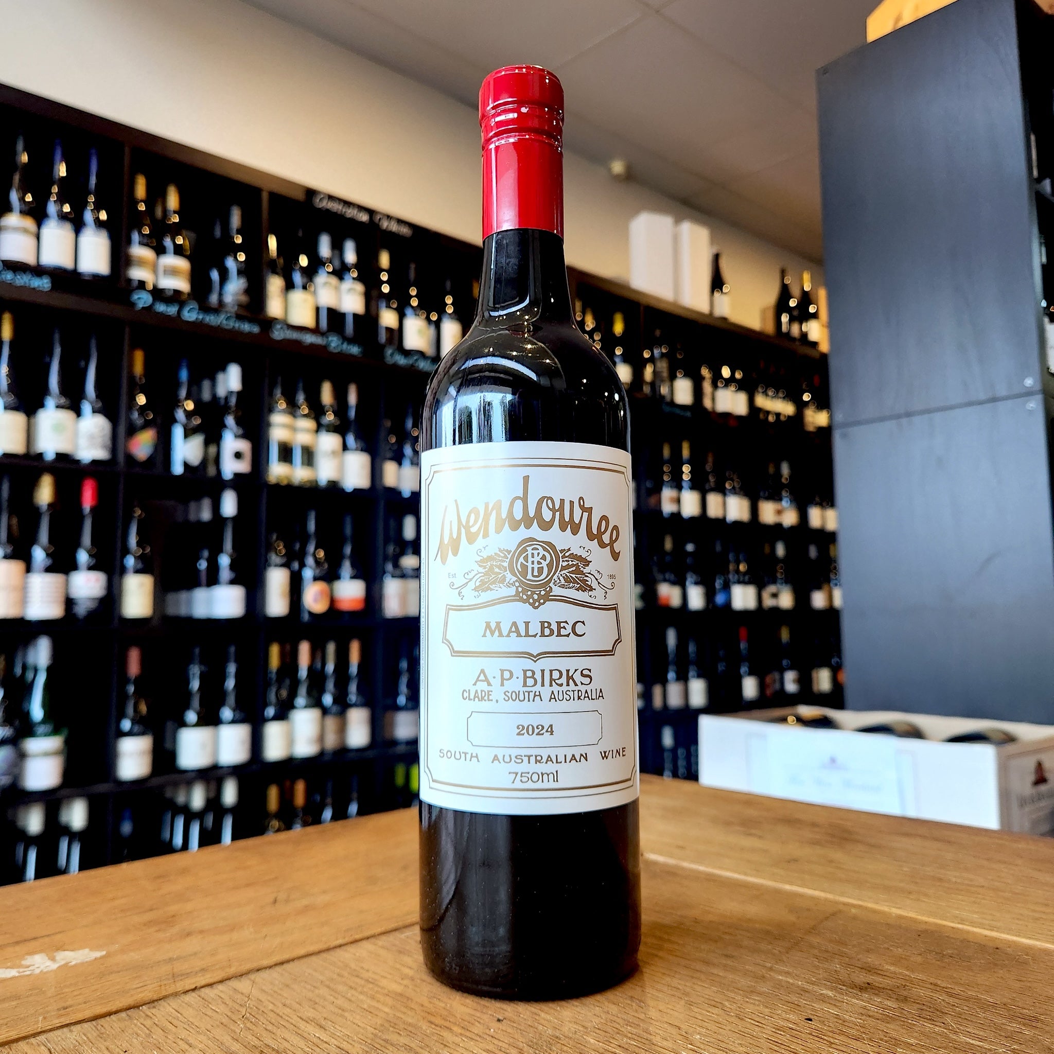 A bottle of Wendouree Wendouree Malbec  stands on a wooden table in a wine shop, with shelves of wine bottles blurred in the background.
