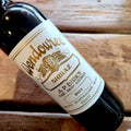 A close-up of a dark glass wine bottle lying on a rustic wooden surface, showing a white label with gold text that reads Wendouree Shiraz, 2024.