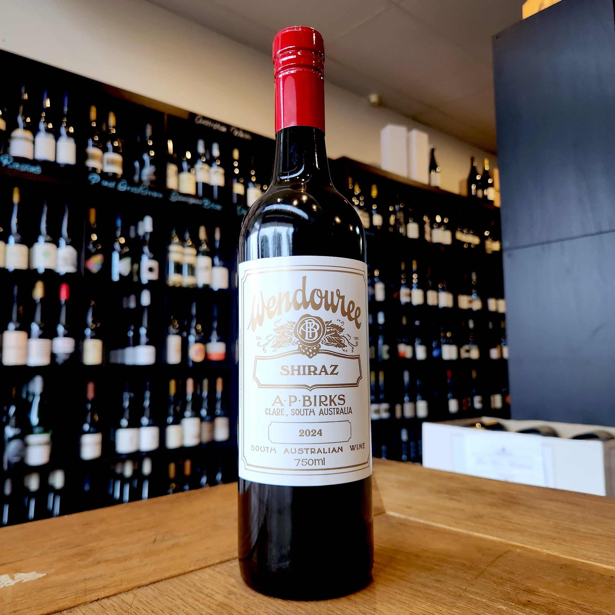 A bottle of Wendouree Shiraz standing on a wooden table in front of a wine shop wall filled with blurred bottles.