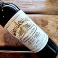 A close-up of a dark glass wine bottle lying on a rustic wooden surface, showing a white label with gold text that reads Wendouree Shiraz Malbec, 2024.