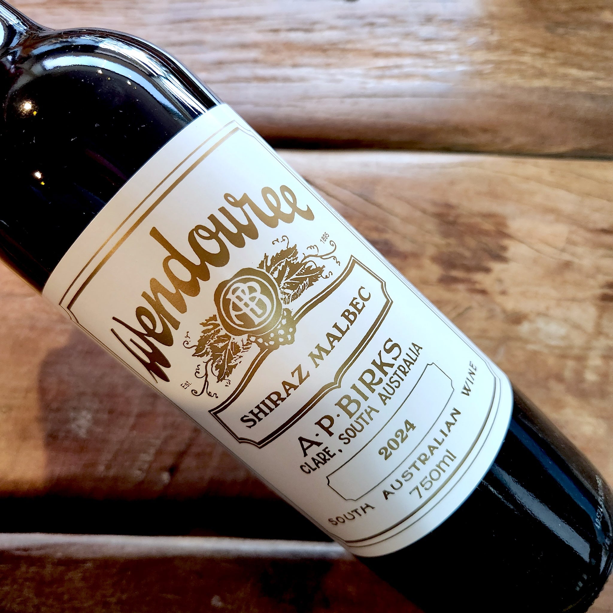 A close-up of a dark glass wine bottle lying on a rustic wooden surface, showing a white label with gold text that reads Wendouree Shiraz Malbec, 2024.