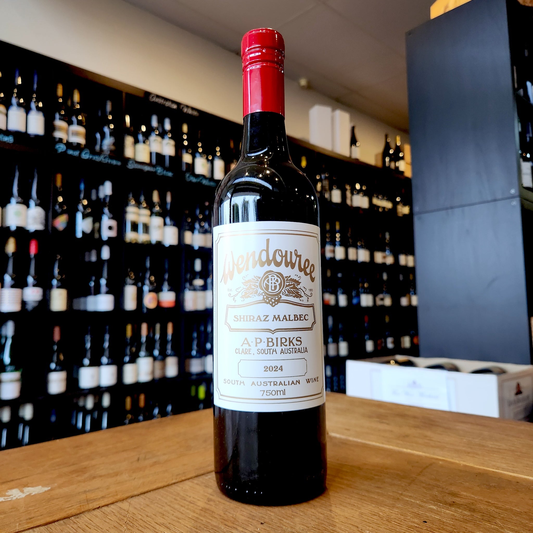 A bottle of Wendouree Shiraz Malbec standing on a wooden table in front of a wine shop wall filled with blurred bottles.