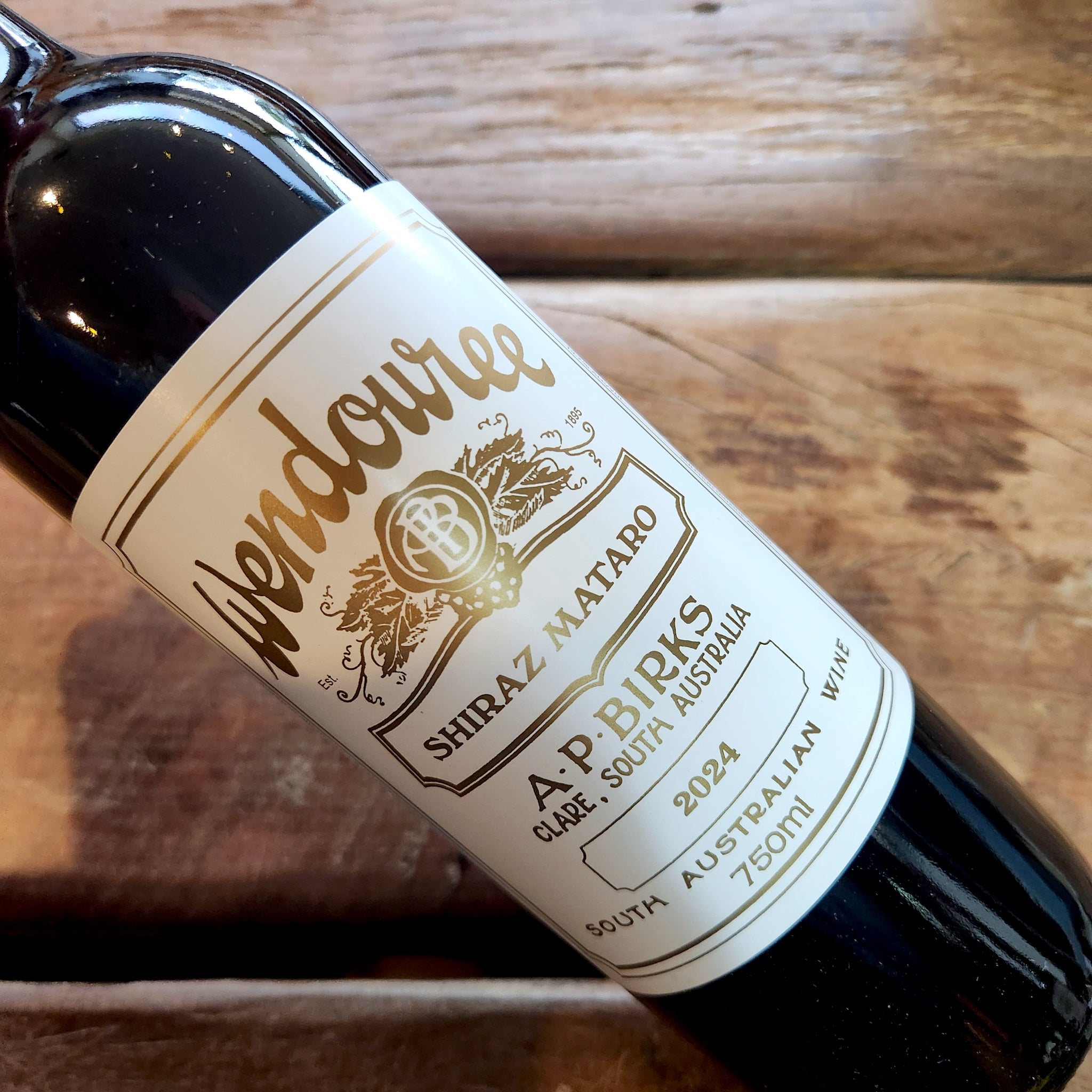 A close-up of a dark glass wine bottle lying on a rustic wooden surface, showing a white label with gold text that reads Wendouree Shiraz Mataro, 2024.
