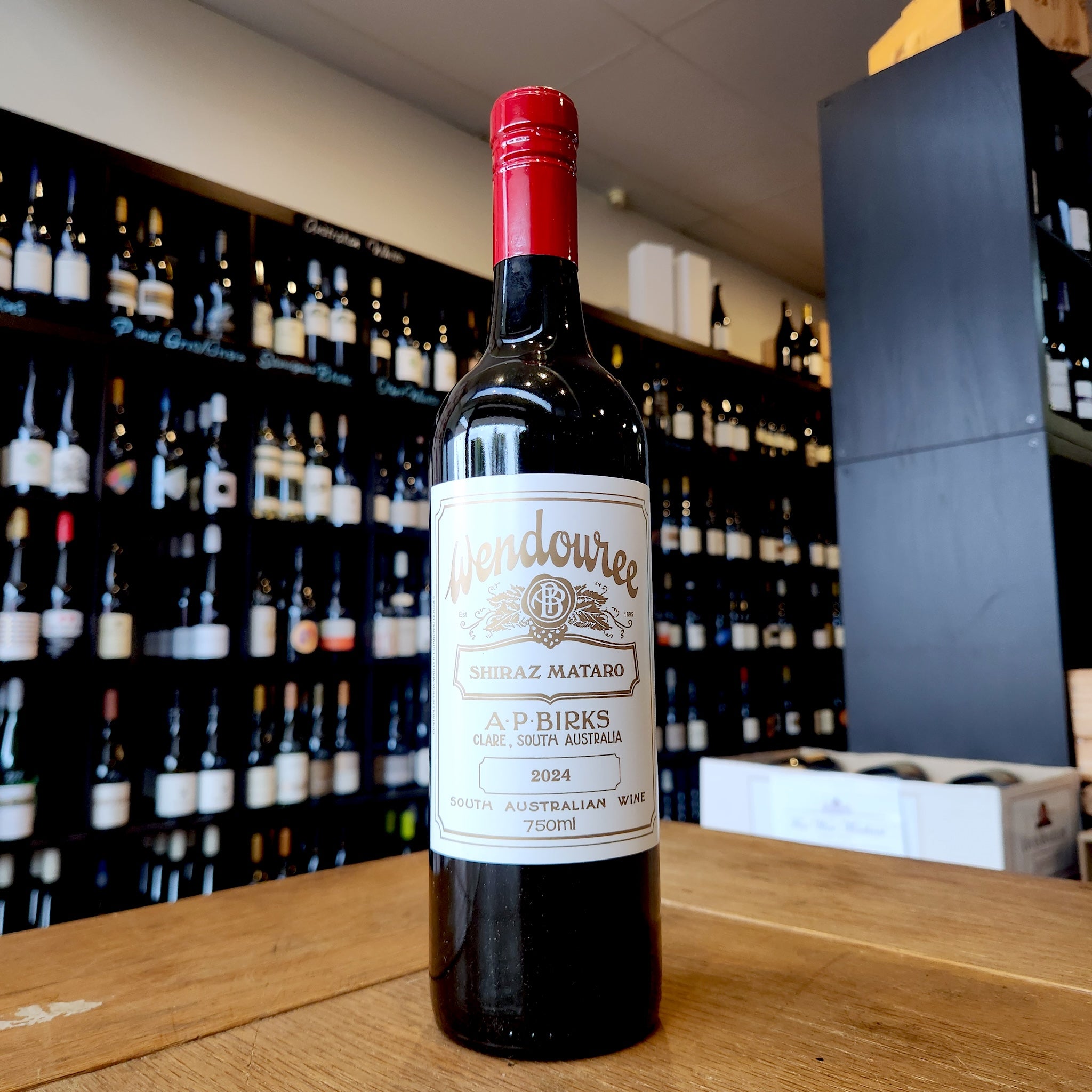 A bottle of Wendouree Shiraz Mataro standing on a wooden table in front of a wine shop wall filled with blurred bottles.